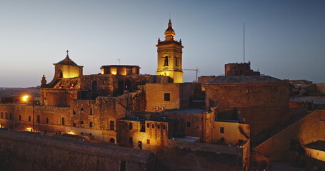 Malta, Comino island: Warm night lights illuminating the Cittadella in Gozo, Malta, highlight the medieval architecture of the fortified city at dusk. Aerial drone flight panorama