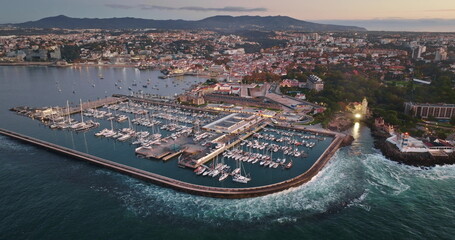 Portugal, Lisbon: Cascais coastal city with vibrant marina bay filled with docked boats yachts, historical buildings, and wide urban landscape extending into mountains at dusk. Aerial view panorama
