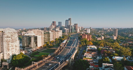 Australia, Sydney: Sydney's skyline at sunrise, skyscrapers, cars driving highway road, residential buildings, and green spaces. Aerial drone flight over modern cityscape, urban architecture