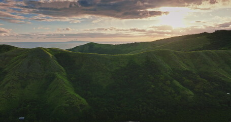 Lush green hills and valleys creating a beautiful landscape in Fiji, with the Pacific Ocean visible in the distance under a dramatic sky with golden sunset light