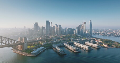 Australia, Sydney: Aerial view of Sydney Harbour Bridge and city skylines skyscrapers reflecting in water at misty sunrise, Barangaroo Reserve and wharves in foreground. Drone flight panorama