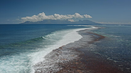 Crystal-clear turquoise water gently washes over a vibrant coral reef. Creating a breathtaking contrast against the deep blue ocean with a mountainous island on the horizon under a bright cloudy sky