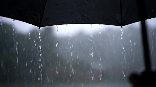 Rainy day scene captured from under a black umbrella, focusing on raindrops falling around it