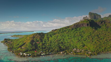 Aerial panoramic view revealing pristine Bora Bora coastline with verdant tropical vegetation. Crystal-clear turquoise lagoon. Traditional houses. Wooden piers nestled in French Polynesian landscape