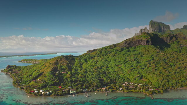 Aerial panoramic view revealing pristine Bora Bora coastline with verdant tropical vegetation. Crystal-clear turquoise lagoon. Traditional houses. Wooden piers nestled in French Polynesian landscape