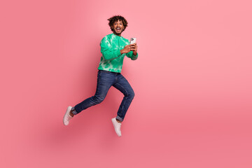 Young man jumps in the air while holding a smartphone against a pink background wearing a green tie...