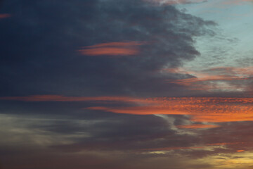 ​A dramatic evening sky is filled with thick dark blue clouds, through which bright orange and golden rays of the setting sun break through.