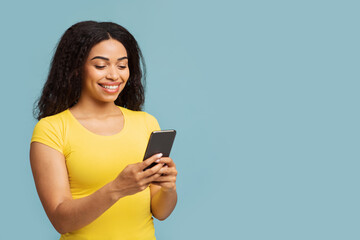 Fototapeta premium Happy african american woman using smartphone texting and networking standing over blue studio background, free space. Black young lady using mobile application on phone