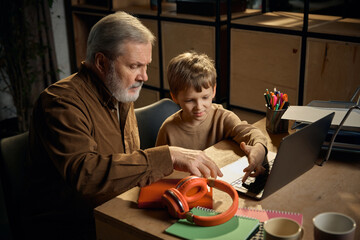 Boy teaching grandfather laptop skills at home workspace. Concept of digital literacy, senior...
