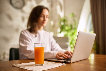 Woman working on laptop with glass of fresh orange juice on wooden table. Concept of healthy remote work routine, balanced lifestyle, hydration and fresh energy during home office day. © Lustre Art Group 