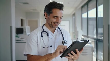 Smiling doctor using tablet in hospital corridor with stethoscope reviewing digital medical records. - Powered by Adobe