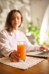 Woman working on laptop with glass of fresh orange juice at home. Concept of healthy remote work routine, daily vitamins, balanced lifestyle, focus, calm productivity and natural energy.