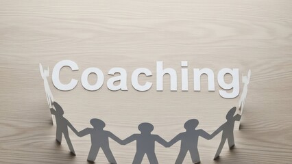 Paper figures hold hands around the word "Coaching" on a wood surface