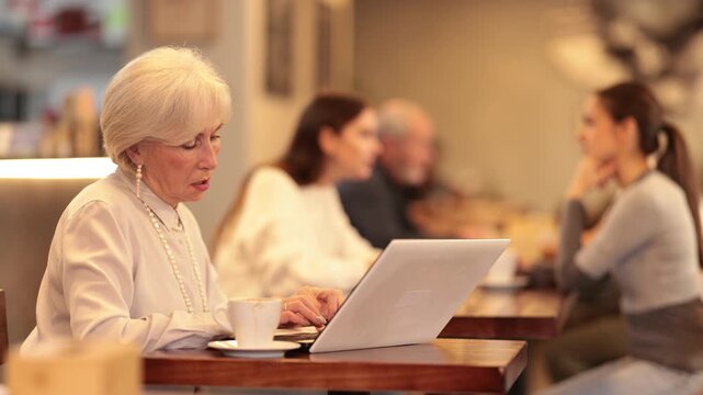 Elderly woman sitting and typing on a laptop in a cozy coffee shop. Adult writer is working on a new book. Remote employee retirees to correspond with a client. High quality 4k footage