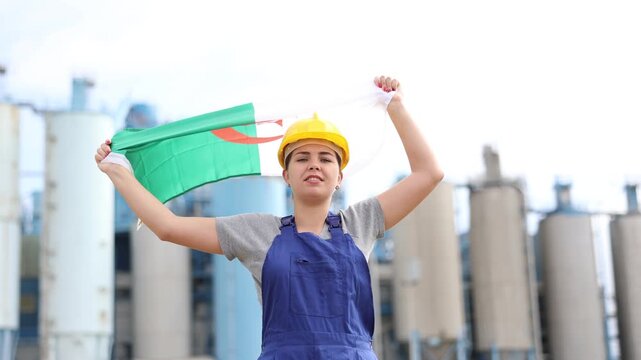 Worried girl in work clothes and hardhat with flag of algeria standing in front of industrial scenery