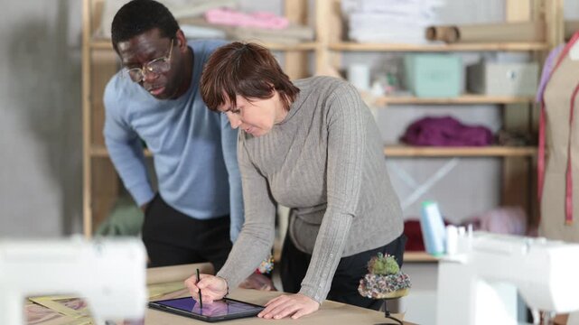 Two interested fashion designers, man and woman, collaborating in sewing studio, using tablet to create and refine new garment design at pattern table, discussing digital sketches or client references