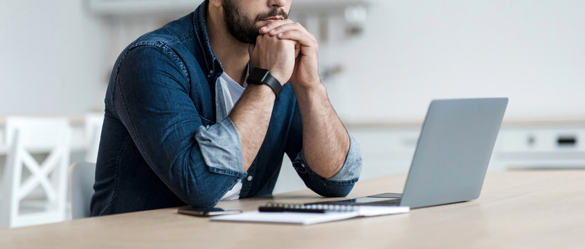 Upset, sad, depression, frustrated and work at home. Concentrated pensive millennial bearded male looks at laptop and pressed hands to mouth, sits at table in kitchen interior, profile, panorama