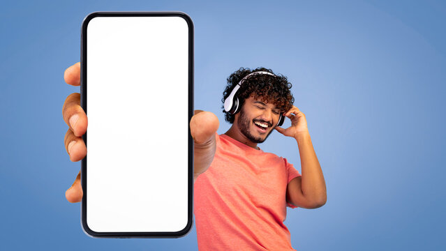 A young man with curly hair enjoys music through headphones. He holds a blank smartphone screen forward while smiling. The background is a simple blue color.