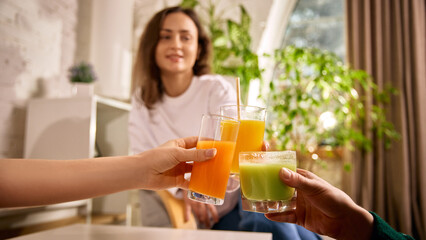 Friends clinking fresh juice glasses at home. Concept of casual meeting, healthy drinks, social bonding, relaxed lifestyle, positive emotions, natural light interior, everyday wellness moment.