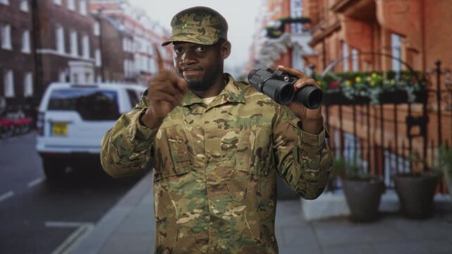 Man soldier in camouflage uniform holding binoculars and pointing finger on a street with parked van and railings; vigilance duty.