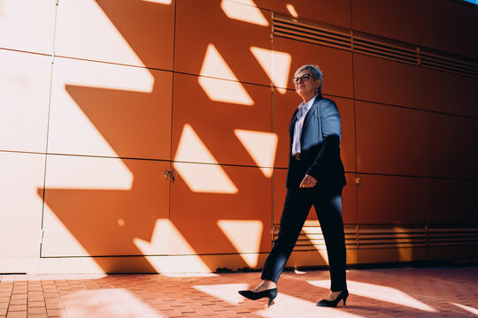 Senior professional woman striding purposefully under dramatic shadows, symbolizing ambition, resilience, and forward motion in a tech-connected urban world.