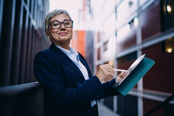 Fototapeta premium Confident senior woman holding stylus and digital tablet, looking at camera with slight smile, symbolizing technological empowerment and modern professional identity in an outdoor space.
