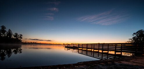 A beautiful sunset over a lake with a pier in the foreground. The water is calm and the sky is filled with clouds
