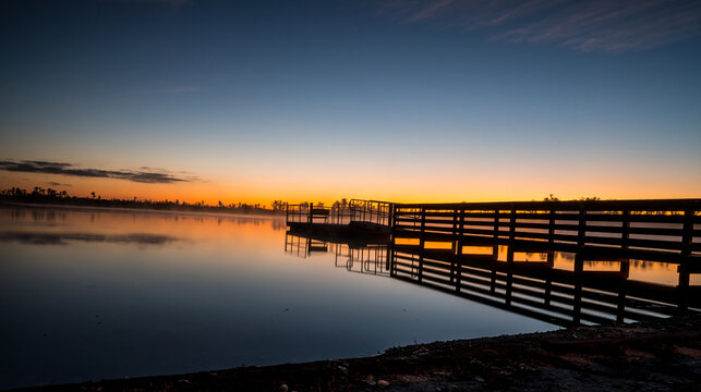 A beautiful sunset over a lake with a pier in the foreground. The water is calm and the sky is a mix of orange and pink hues