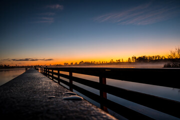Obraz premium A wooden bridge over a body of water with a beautiful sunset in the background. The water is calm and the sky is filled with orange and pink hues. The scene is serene and peaceful