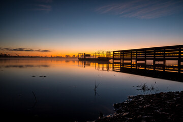 A dock is seen in the water with the sun setting in the background. The water is calm and the sky is a beautiful orange color