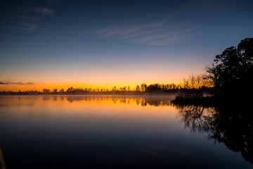 Obraz premium A beautiful sunset over a lake with trees in the background. The sky is a mix of orange and blue hues, creating a serene and peaceful atmosphere. The water is calm and still