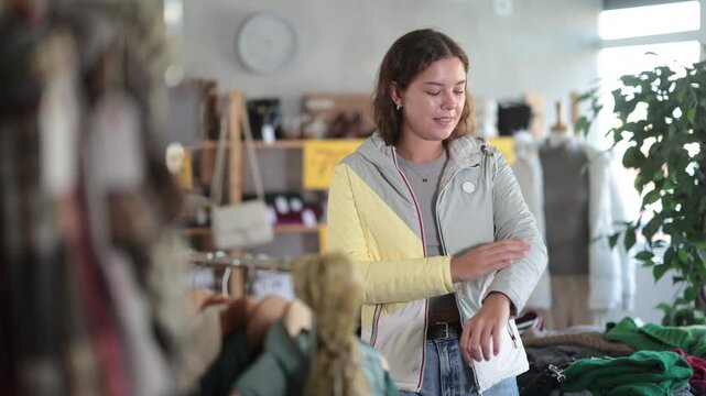  Fashionista girl in shop, go for fitting down jacket. Off line clothing store from retailer. Style and fashion type look, modern trends. Outlet, discount store. Customer looks at herself in mirror