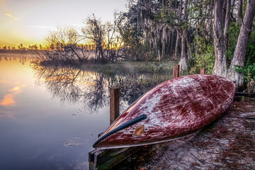 A red canoe sits on the shore of a lake. The sun is setting, casting a warm glow over the scene. The water is calm and still, reflecting the beautiful colors of the sky