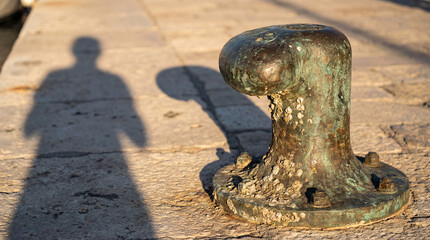 An aged, weathered mooring bitt, speckled with barnacles, stands firmly on the stone quay in Murano. The setting sun creates a ghostly shadow of a person nearby.