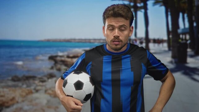 Man holding soccer ball, rubs eyes with forearm on sunlit street promenade by the seaside; fatigue contemplation.