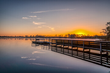 Fototapeta premium A dock is on the water with a sunset in the background. The water is calm and the sky is orange