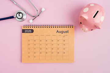 August 2026 desk calendar and stethoscope with piggy bank. Savings for Health concept.