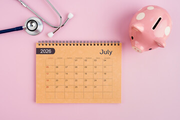 July 2026 desk calendar and stethoscope with piggy bank. Savings for Health concept.