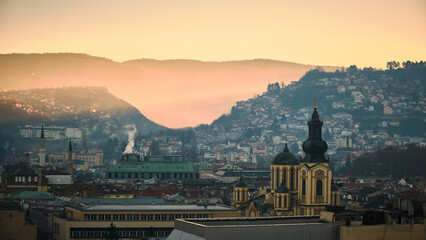 View of Cathedral of the Nativity of the Theotokos rising above Sarajevo buildings in soft morning haze. Hills in background fade into pale atmospheric tones, adding depth and calm, cinematic mood