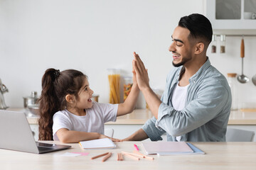 Handsome arab man single father helping his cute little daughter with homework, sitting together in front of laptop at kitchen, giving each other high five, copy space. E-education, home schooling
