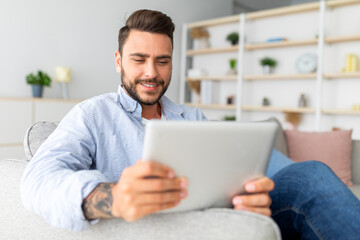 Closeup of caucasian man resting on sofa with digital tablet, home interior, free space. Happy guy sitting on couch in living room, using nice mobile app on modern pad, copy space