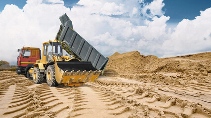 Heavy machinery works on sandy ground leaving deep tracks as a large pile of sand rises in the background under a cloudy sky at a construction site