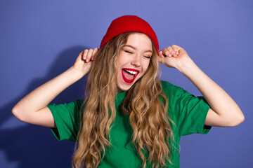 Joyful young woman in green shirt and red beanie dancing against a purple background smiling widely