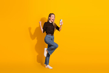 Young woman in casual jeans and brown shirt jumps for joy while taking a selfie against a bright...