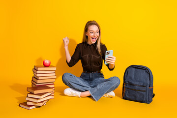 Young woman student celebrates success while posing with phone near a stack of books and a backpack...
