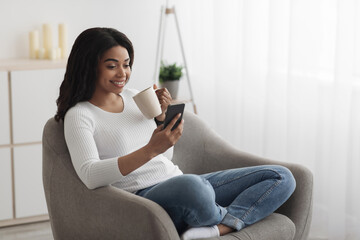 Leisure time. Happy african american lady using smartphone, sitting in armchair and drinking coffee, enjoying morning, free space. Lady surfing internet or messaging online on phone