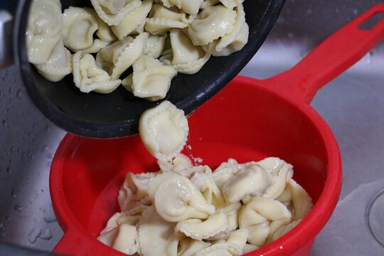 Freshly cooked tortellini are poured from the pot into a red plastic sieve that is standing in the sink