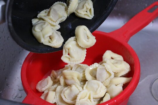 Freshly cooked tortellini are poured from the pot into a red plastic sieve that is standing in the sink