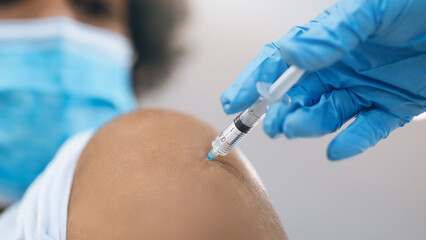 A healthcare worker administers a vaccine to a patient in a medical facility. The patient has a bandage on their arm and wears a face mask. The worker is wearing gloves and a mask.