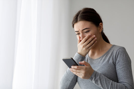 Upset depressed millennial pretty woman covering her mouth with hand and looking at phone, got bad news at home. Human face expression, negative emotion, breaking up with boyfriend during covid-19